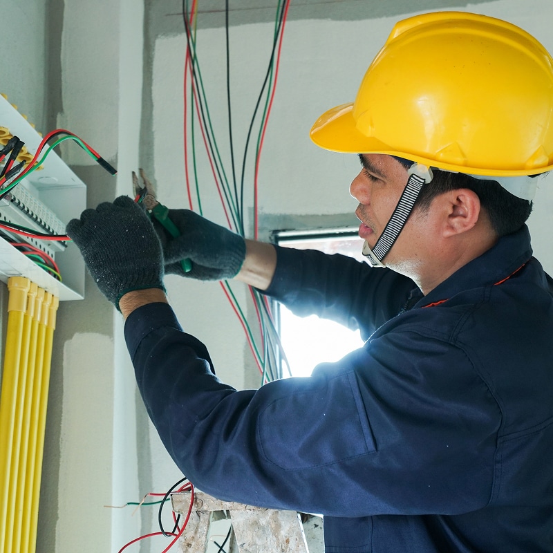 Électricien concentré en casque jaune et gants, câblant un panneau électrique avec des fils colorés sur un chantier.