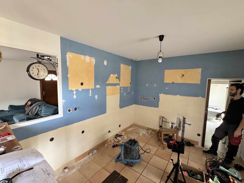 Travaux de rénovation cuisine : murs bleus et crème en cours, outils, aspirateur, plan et homme regardant l'avancement. Horloge murale visible.
