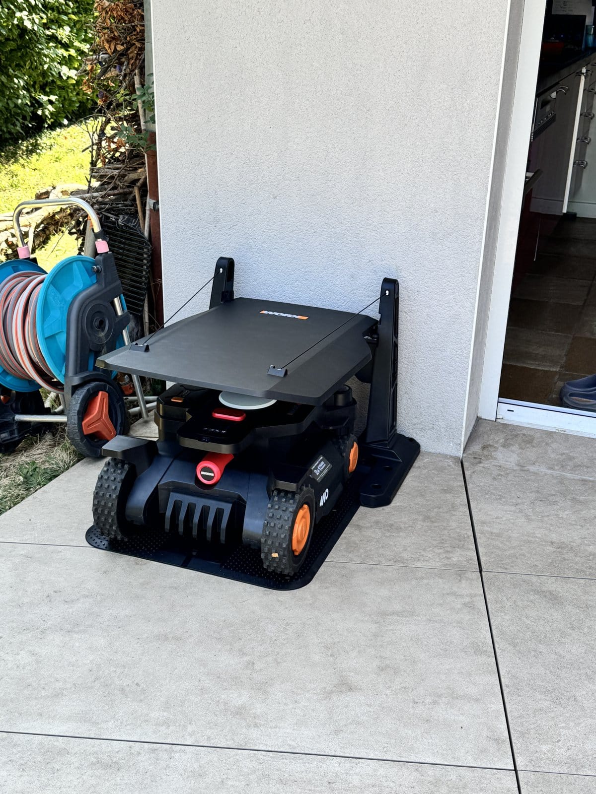 Tondeuse robot WORX Vision noire dans sa station de charge avec abri de protection sur une terrasse carrelée.
